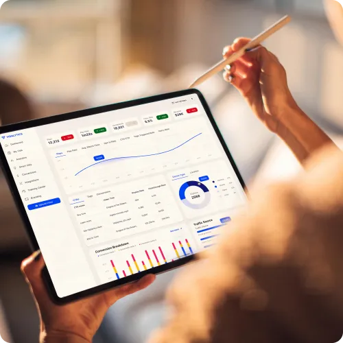 Hands holding a tablet displaying a business analytics dashboard with graphs, charts, and metrics. A pencil is held above the screen.